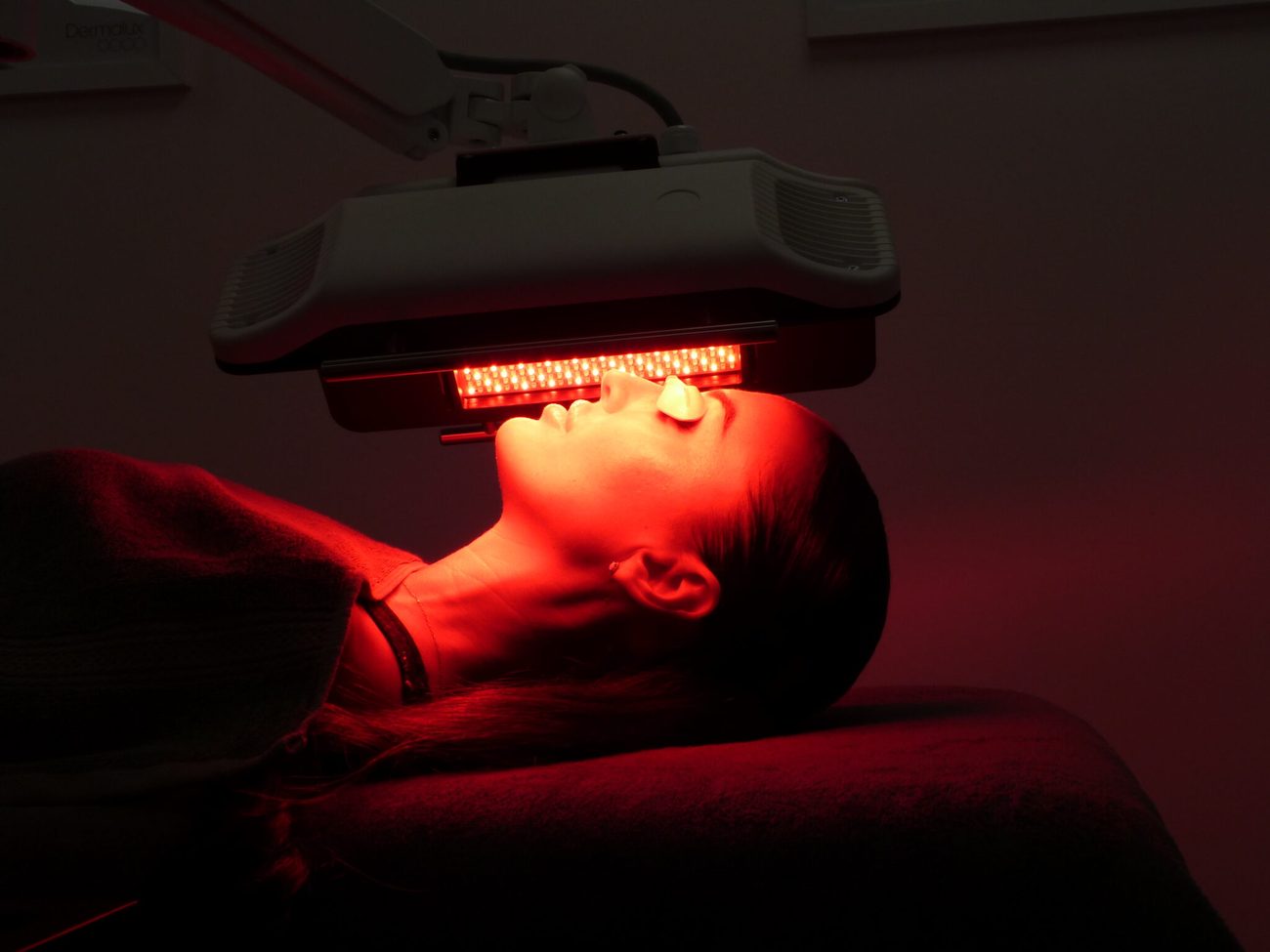 A red LED phototherapy canopy positioned over a client's face during a treatment